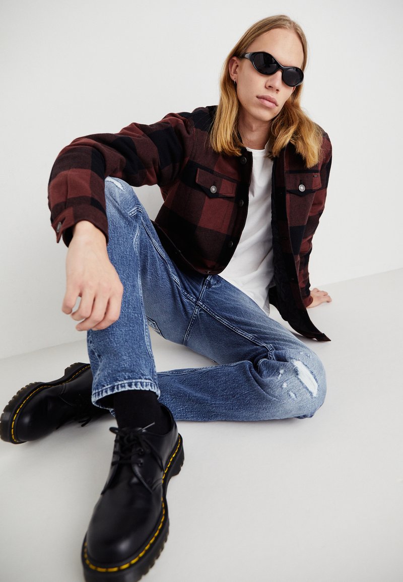 Flanellen jas in rood en zwart ruitpatroon, witte t-shirt, versleten blauwe jeans en zwarte veterschoenen met gele zoolaccenten.