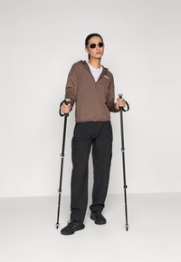 Giacca a zip marrone, t-shirt bianca, pantaloni cargo neri, scarpe nere. Tiene bastoni da trekking; indossa occhiali da sole. Abbigliamento outdoor semplice e funzionale.