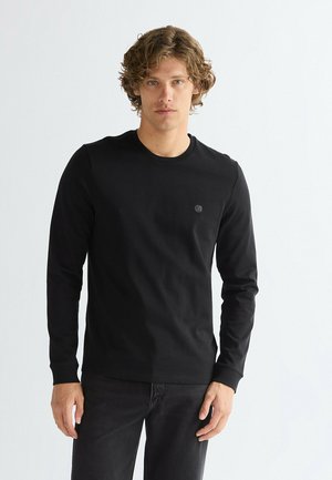 Giovane uomo dai capelli ricci che indossa una maglia nera aderente a maniche lunghe con un piccolo logo ricamato su sfondo bianco.