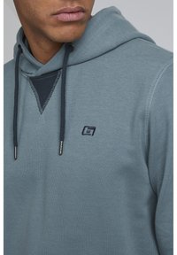 Sudadera con capucha de suave tejido azul texturizado. Presenta un acento de color azul oscuro en el cuello, cordones ajustables y un pequeño logo.