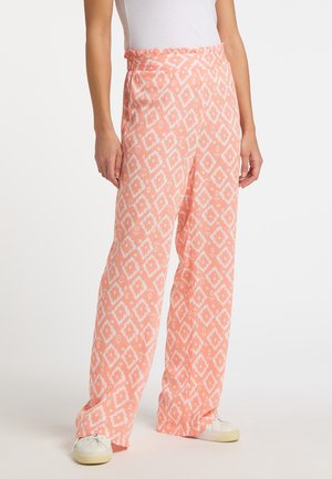 Vrouw draagt losse koraalkleurige broek met witte diamantpatronen en een wit mouwloos topje, gecombineerd met witte sneakers.