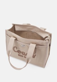 Sac cabas beige en toile Coach New York avec double poignée, poche devant à pression, et ouverture supérieure montrant l’étiquette en tissu à l’intérieur.