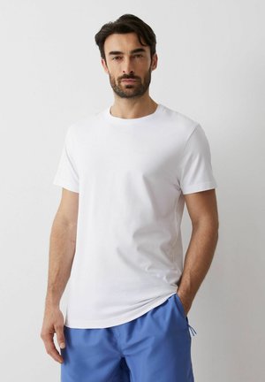 Mężczyzna z brodą, ubrany w gładki biały T-shirt z krótkim rękawem i niebieskie krótkie spodenki, stoi na jasnoszarym tle.