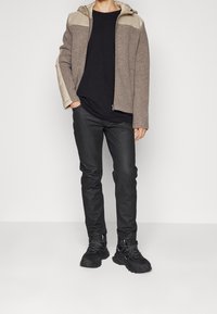 Giacca marrone chiaro con sezioni superiori beige e cappuccio, abbinata a una t-shirt nera, pantaloni neri e scarpe da ginnastica nere. Texture liscia, design casual.