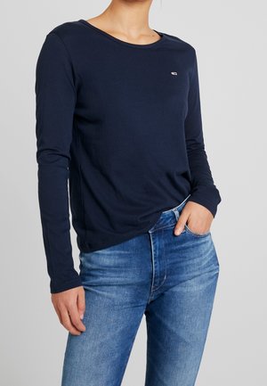 Vrouw draagt een marineblauw shirt met lange mouwen en blauwe jeans, hand in de zak, tegen een effen lichtgrijze achtergrond.