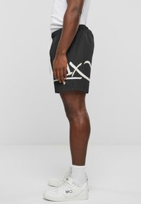 Sorte vævede shorts med elastisk talje, prydet med hvide grafiske accenter. Kombineret med hvide sneakers og ankelsokker.