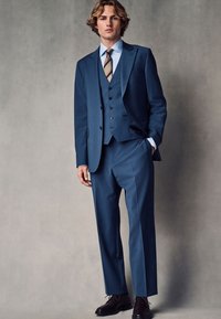 Jeune homme portant un costume trois pièces bleu sur mesure avec une cravate rayée et des chaussures noires, debout devant un fond gris uni.