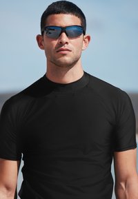 Schwarzes Rash-Guard mit kurzen Ärmeln aus dehnbarem, glattem Material, kombiniert mit dunklen, reflektierenden Sonnenbrillen. Nahaufnahme des Oberkörpers.