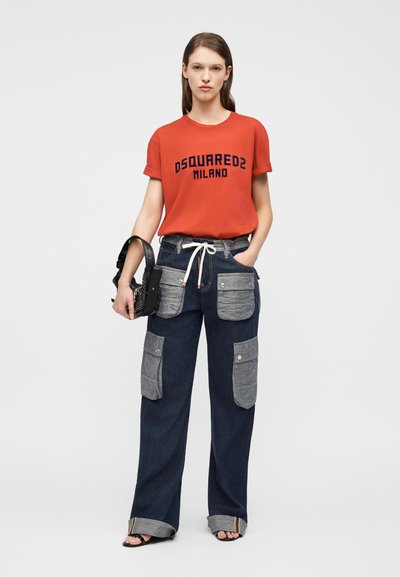 Γυναίκα στέκεται με πρόσωπο προς τα εμπρός φορώντας πορτοκαλί μπλούζα DSQUARED2 Milano, σκούρο τζιν με μεγάλες γκρι τσέπες, μαύρα σανδάλια και κρατώντας μια μαύρη τσάντα με τρουκς.
