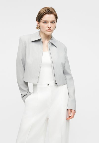 Νέα γυναίκα με κοντά μαλλιά, που φοράει ένα ανοιχτό γκρι κοντό μπουφάν πάνω από ένα λευκό τοπ και ψηλόμεσα λευκά παντελόνια, στέκεται με το ένα χέρι στην τσέπη.