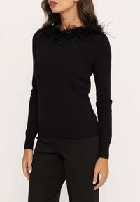 Zwarte trui met lange mouwen en een veren zwarte kraag. Zacht gebreid materiaal met een aansluitende pasvorm. Gecombineerd met zwarte broeken.