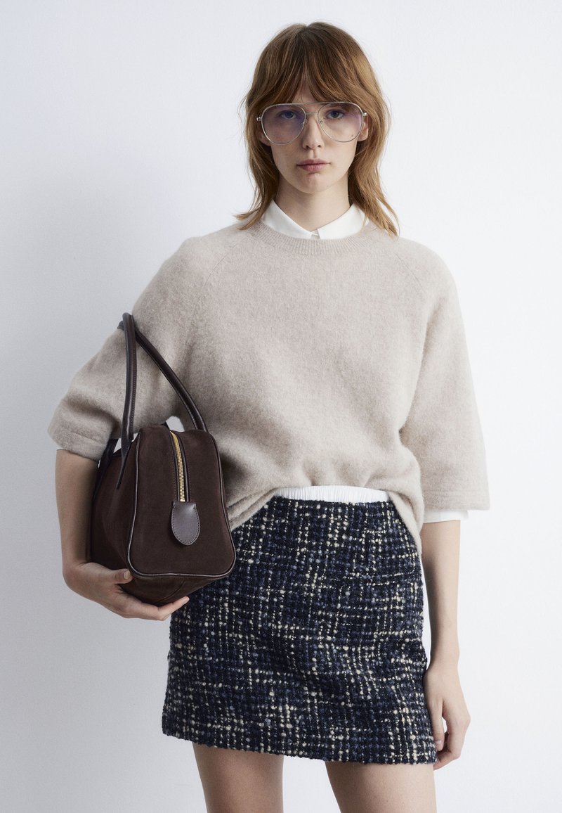 Pull en beige duveteux, chemise blanche à col en dessous, jupe texturée bleu marine et crème, et sac à main marron foncé avec fermeture éclair.