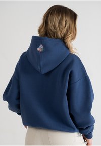 Felpa blu navy con cappuccio e vestibilità ampia; presenta un piccolo dettaglio di ricamo floreale colorato sul retro vicino al collo.