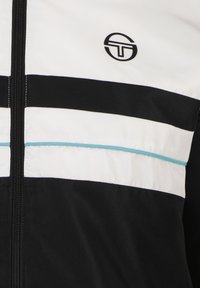 Zwart-witte zip-up jas met een textuurafwerking, horizontale strepen en een turkoois accentlijn. Logo gepositioneerd op de borst.