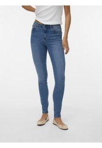 Femme portant un jean skinny bleu clair, un haut blanc et des ballerines beiges, debout sur un fond clair uni.