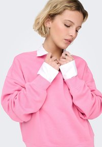 Ροζ φούτερ με στρογγυλή λαιμόκοψη, ευρύχωτη εφαρμογή και ριμπ μανσέτες. Λευκό πουκάμισο με γιακά φαίνεται κάτω από το φούτερ, εμφανές στον γιακά και τις μανσέτες.