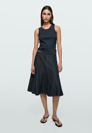 Femme portant un haut noir sans manches, une jupe trapèze en denim sombre et des tongs noires, debout devant un fond clair uni.