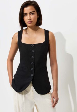 Giovane donna con capelli corti e scuri indossa un gilet nero senza maniche abbottonato e pantaloni larghi chiari su uno sfondo semplice.