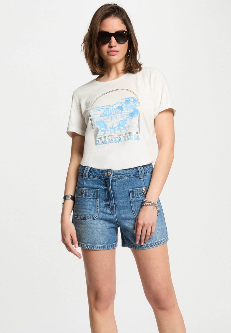 Maglietta bianca in cotone con design grafico blu, abbinata a pantaloni corti in denim azzurro chiaro con tasche anteriori e bottoni decorativi. Occhiali da sole neri.
