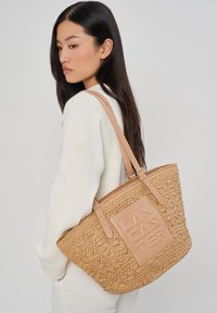 Femme aux longs cheveux foncés portant un pull blanc, avec un grand sac fourre-tout tressé de couleur beige, orné d'un écusson logo embossé "LANCEL" sur l'épaule.