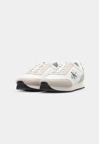 Tenisky Calvin Klein s bílým a béžovým semišovým svrškem, černou gumovou podrážkou, šněrováním v přední části a logem na boku. Obsahují prvky retro designu.