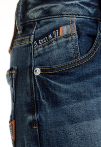 Primer plano de un bolsillo de jeans de mezclilla azul oscuro con costuras naranjas, un remache de metal y una etiqueta de tela que dice "9 CUSTOM 97".