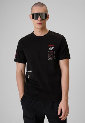 Dorosła osoba ubrana w czarny t-shirt z grafiką i czarne spodnie, z dużymi czarnymi okularami przeciwsłonecznymi z daszkiem, stojąca na jednolitym szarym tle.
