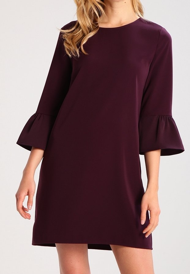 Kvinna klädd i en knälång, mörklila klänning med 3/4-ärmar och rund halsringning framför en enkel ljus bakgrund.