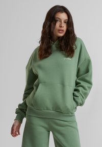 Haut à capuche vert surdimensionné avec une poche kangourou et des poignets côtelés. Présente l'inscription "Interdit" brodée sur la poitrine. Fabriqué en tissu doux.