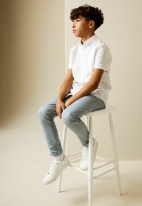 Camisa blanca de manga corta, pantalones de mezclilla azul claro y zapatillas blancas con suelas de goma texturizadas. Sentado en un taburete blanco frente a un fondo neutro.