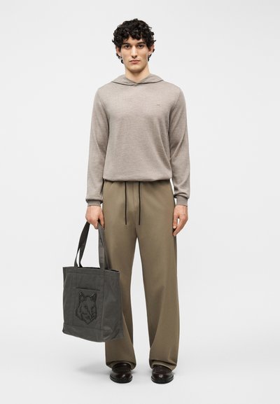 Άντρας που στέκεται, φορώντας μπεζ φούτερ με κουκούλα και ελιά παντελόνι, κρατώντας μια σκούρα γκρι τσάντα ώμου με σχέδιο κεφαλής λύκου.
