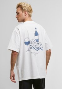 Hvit bomull t-skjorte med løs passform, med et blått grafisk trykk av vin, ost, kjeks og et skjærebrett på baksiden.