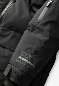 Manche d'une veste en duvet noire avec une poche zippée, un poignet en velcro et des détails de référence imprimés avec des lignes et des coordonnées. Texture lisse.