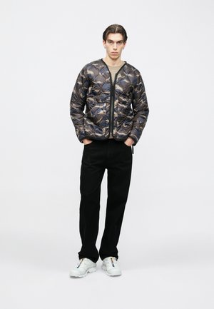 Young man standing with hands in pockets, wearing a blue and beige patterned quilted jacket, black pants, and white sneakers with colorful details.