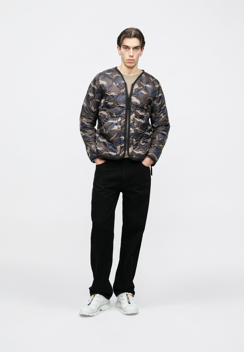 Fiatal férfi zsebében tartott kézzel, kék-beige mintás steppelt dzsekit, fekete nadrágot és színes részletekkel ellátott fehér tornacipőt viselve áll.