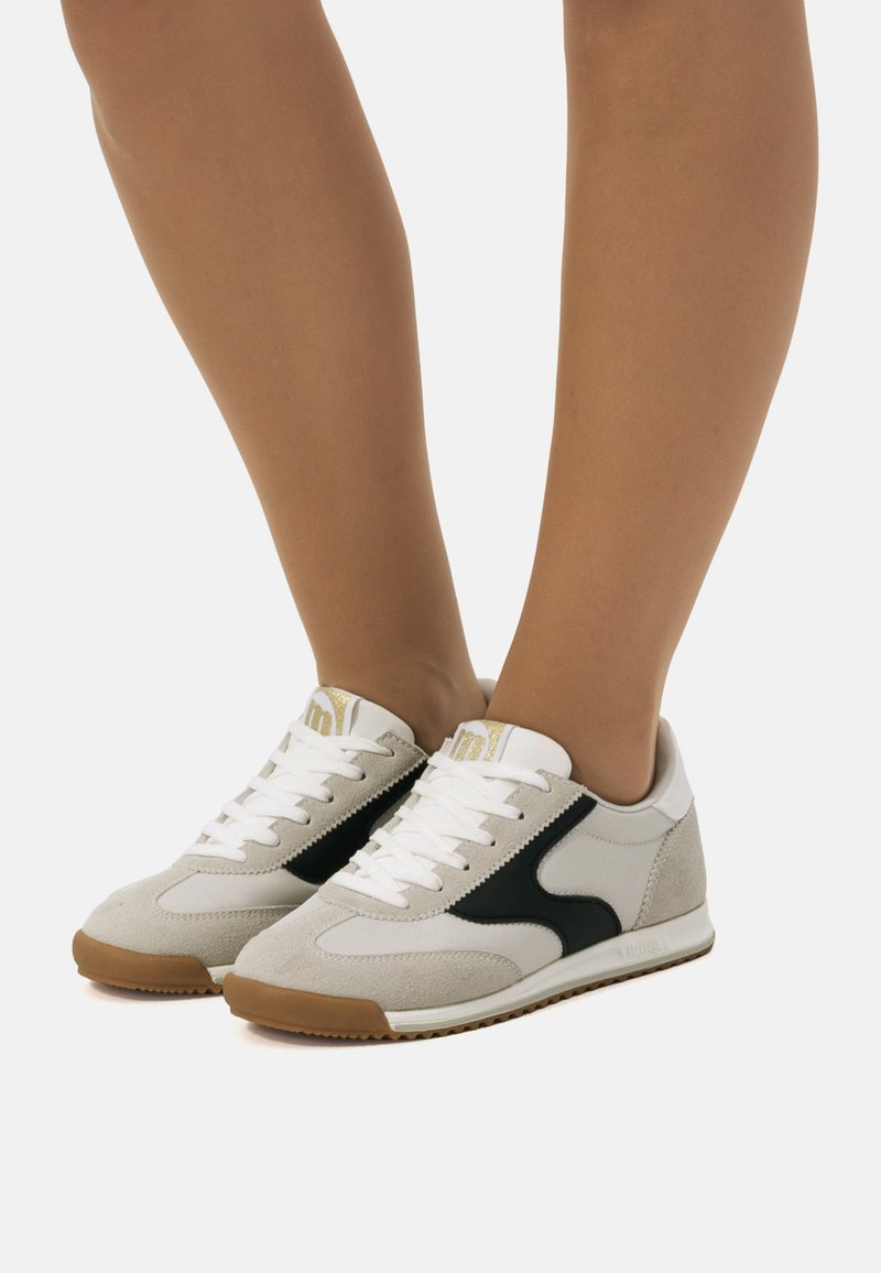Leichte Sneaker aus weißem und schwarzem Wildleder, mit einem strukturierten Lederoberteil, weißen Schnürsenkeln und einer Sohle aus Gummi. Goldene Akzente auf der Zunge.