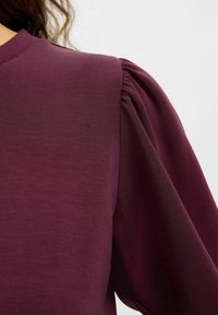 Burgundská bluzka s hladkým povrchem, která má nabírané balónové rukávy a kulatý výstřih. Materiál vypadá lehce a jemně.