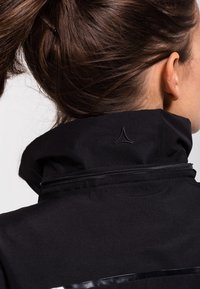 Svart jacka med hög krage, texturerat tyg och en liten logodetalj på ryggen. Dragkedja längs kragen.