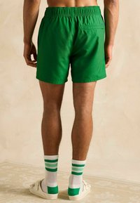 Bărbat îmbrăcat cu șorturi verzi, șosete albe cu dungi verzi și sandale slip-on albe, stând pe un podea bej cu un model, în fața unui zid simplu.