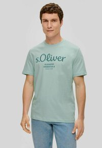 Hellgrünes Baumwoll-T-Shirt mit kurzen Ärmeln, auf der Vorderseite mit einem dunkelblauen Logo und Text versehen. Getragen mit hellblauen Jeans.