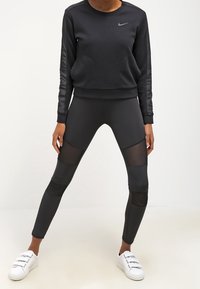 Zwarte Nike-sweater met textuursleeves, gecombineerd met zwarte legging met meshpanelen. Draagt witte sneakers.