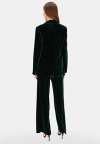 Zielony aksamitny garnitur w kolorze szmaragdowym z dopasowaną marynarką i szerokimi spodniami, charakteryzujący się gładką teksturą i czystą, uporządkowaną sylwetką.