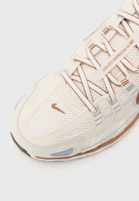 Nike tenisica s gornjim dijelom od kremaste mreže, srebrnim akcentima, smešim šavovima i gumenenim potplatom. Ima vezice i mali logotip sa strane.