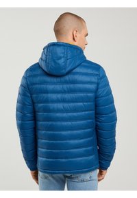 Egy férfi háttal, kék steppelt kapucnis dzsekit és világoskék farmert visel, egyszínű, világos háttér előtt.