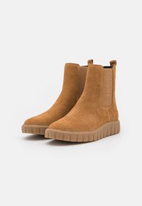 Brązowe zamszowe botki za kostkę z elastycznymi panelami bocznymi, teksturowaną gumową podeszwą i akcentem z logo po boku. Płaski obcas, nowoczesny design.