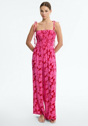 Mujer de pie con las manos detrás de la espalda vistiendo un mono sin tirantes con estampado floral en rosa y rojo y sandalias con tiras transparentes sobre fondo blanco.