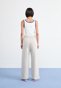 Person med kort mörkt hår som bär en vit linne med marinblå kant, beige vida byxor och sneakers står på en blått golv vänd bort.