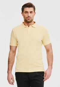 Żółta koszulka polo z klasycznym kołnierzykiem i krótkimi rękawami. Posiada centralną listwę guzikową oraz subtelną logo na klatce piersiowej.