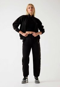 Black athletic tracksuit featuring a zip-up jacket with elastic cuffs and matching pants with side stripes and drawstring waist. Finished with sneakers.