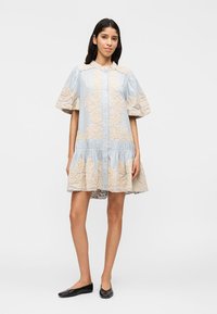 Γυναίκα που φοράει ένα ρούχο με μπροστινά κουμπιά σε ανοιχτό γαλάζιο χρώμα, με μπεζ κεντήματα με λουλούδια και φουσκωτά κοντά μανίκια, σε συνδυασμό με μαύρα μπαλαρίνες.